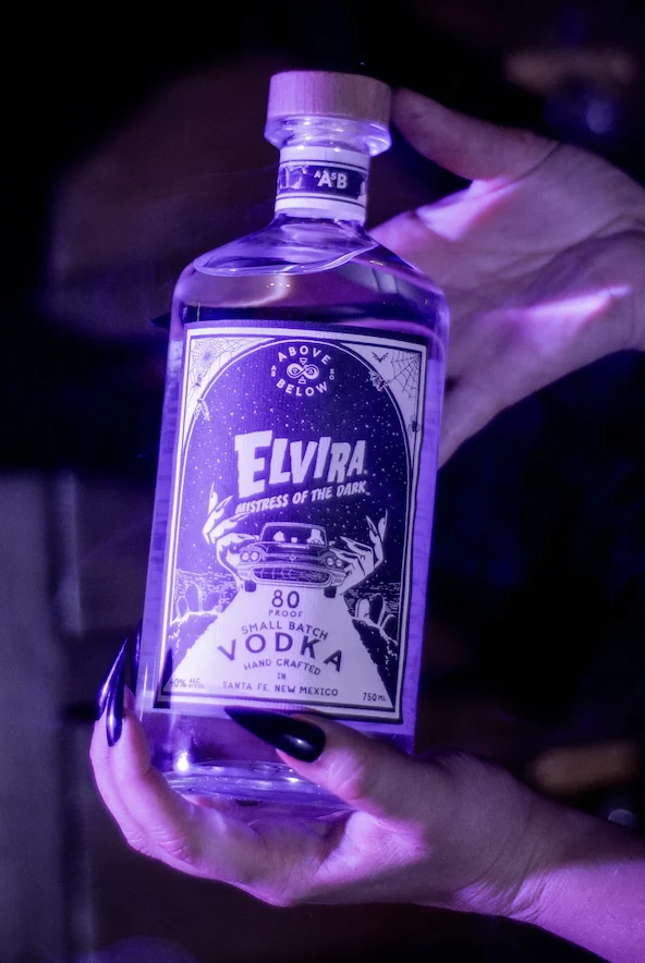 Hands holding a bottle of Elvira, Mistress of the Dark Small Batch Vodka, lit in purple